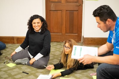 Magaly Urdiales smiles while sitting on the floor next to two people in a work group.